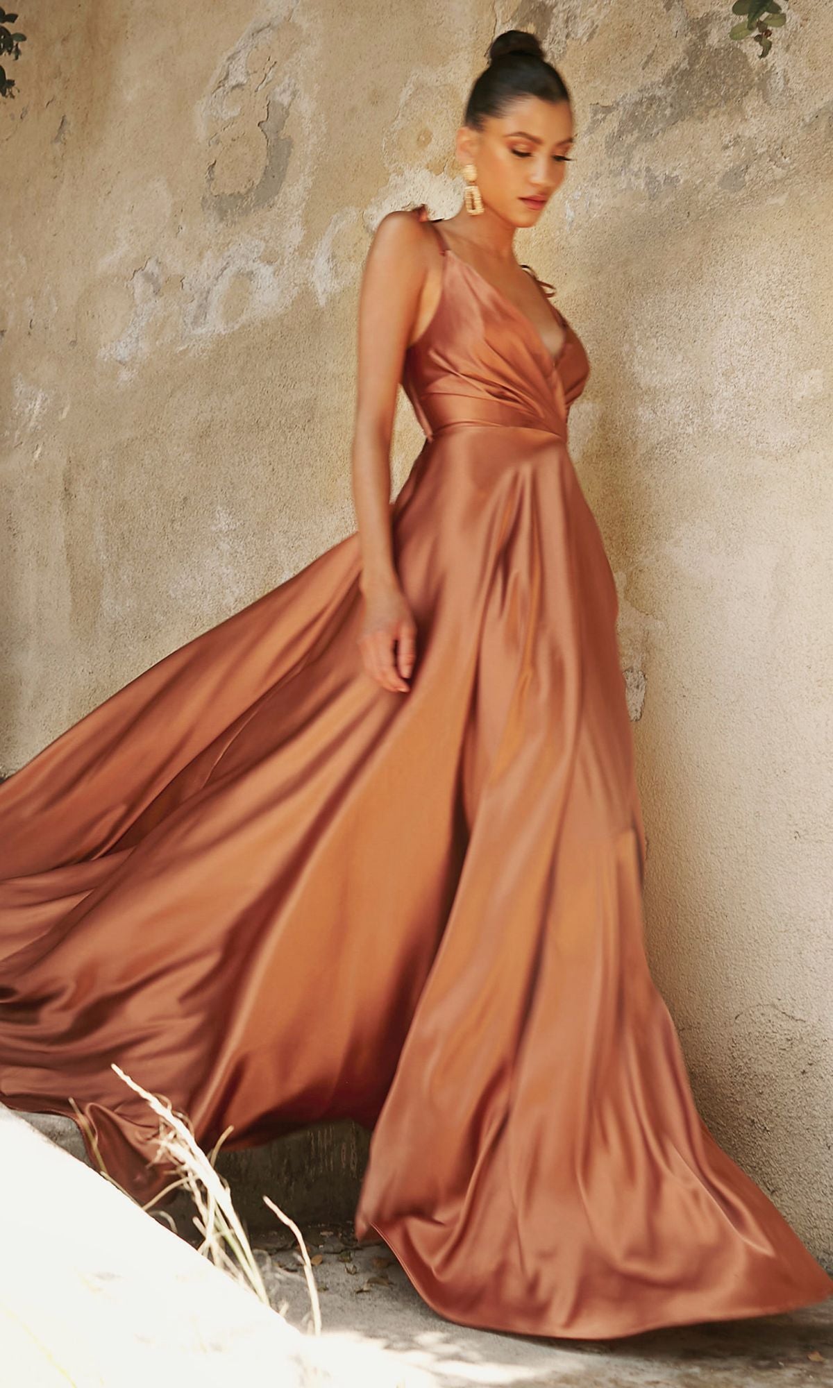 A woman wearing the Shoulder-Tie Long Satin V-Neck Prom Dress BD105 in a copper hue stands before a textured beige wall, her hair styled elegantly in a high bun.